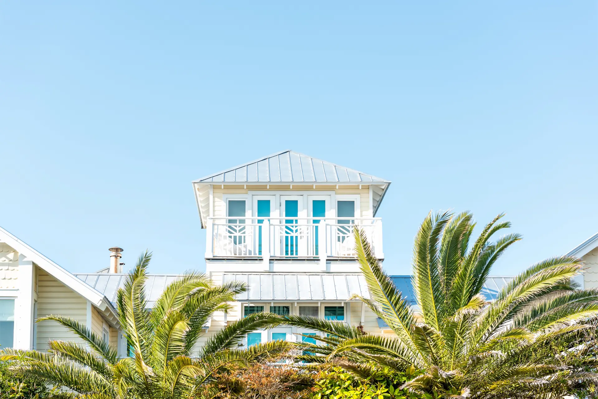 Elevated white coastal Florida beach house with a metal roof and balcony framed by palm fronds under a clear blue sky in Manatee County